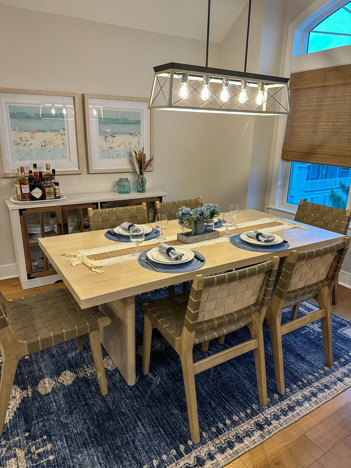 Coastal dining room with woven leather chairs, blue patterned rug, and beach artwork