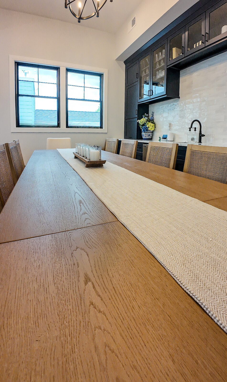 Long white oak dining table with cane back chairs and a dark built-in bar wall behind