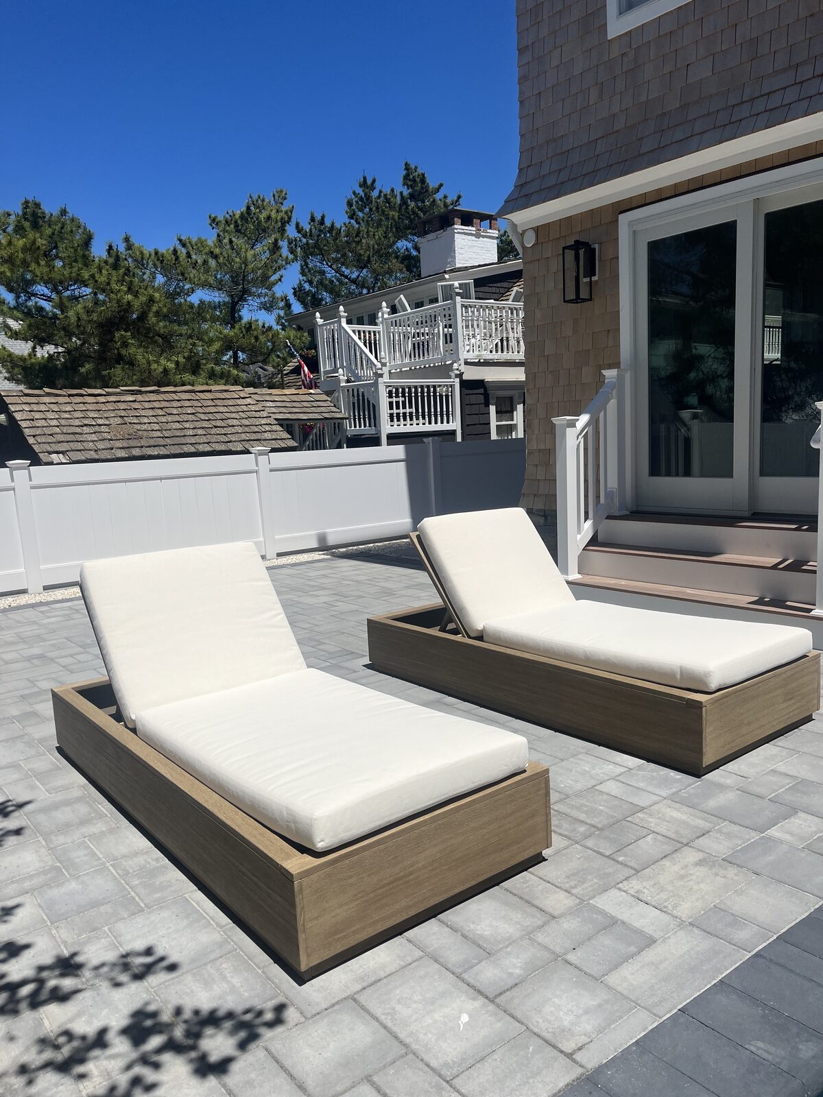 Teak chaise lounges with white cushions on a paver patio outside a shingled beach home